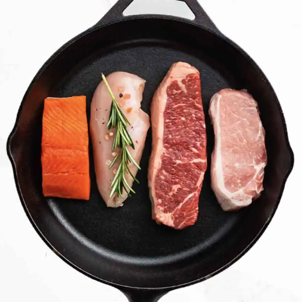 Assorted raw meats including steak, chicken, and salmon on a black cast iron skillet.