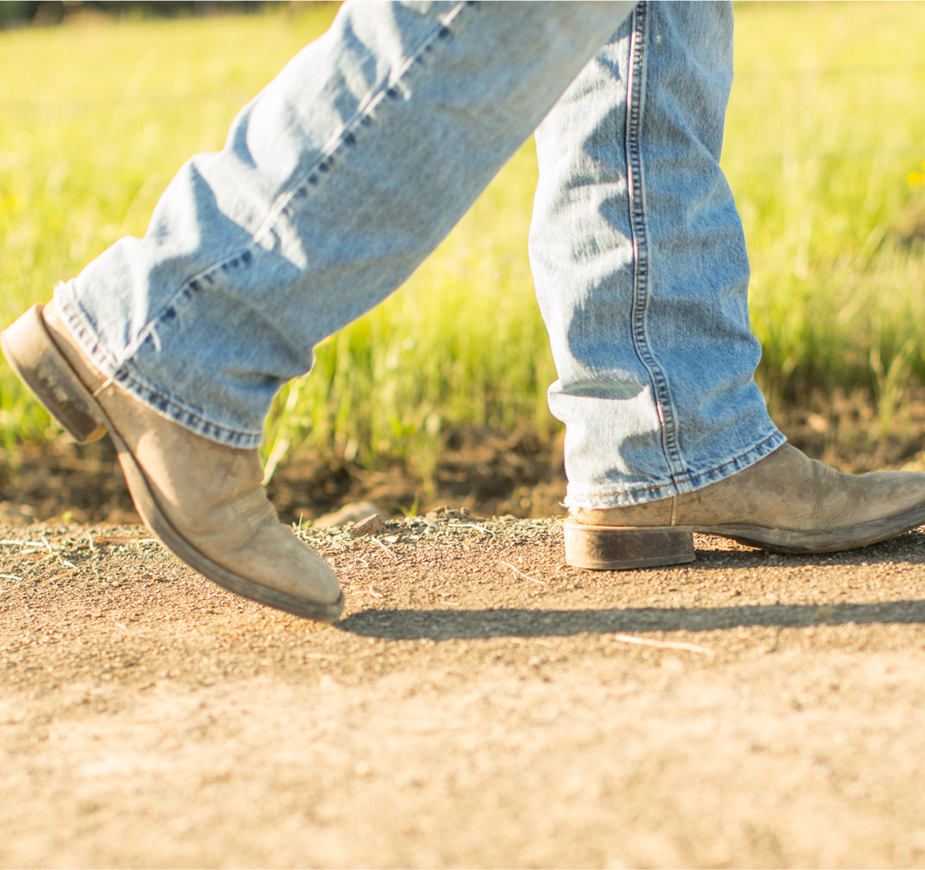 Close-up of legs wearing blue jeans and brown boots walking on a dirt path with grass in the background.
