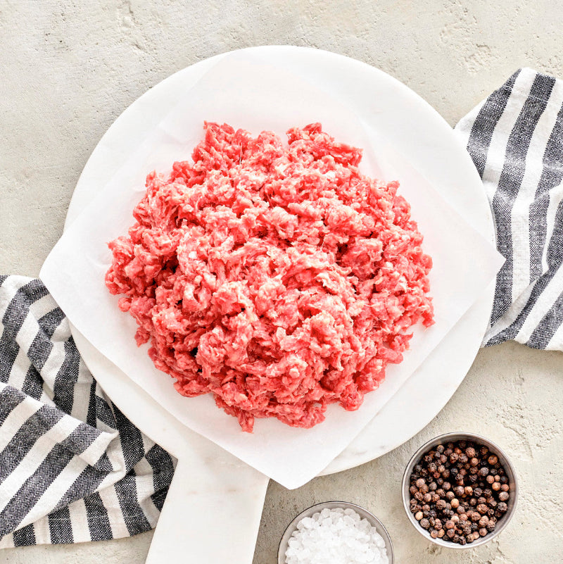 Raw ground beef on a white plate with ingredients for cooking, such as salt and pepper, and a striped napkin in the background.