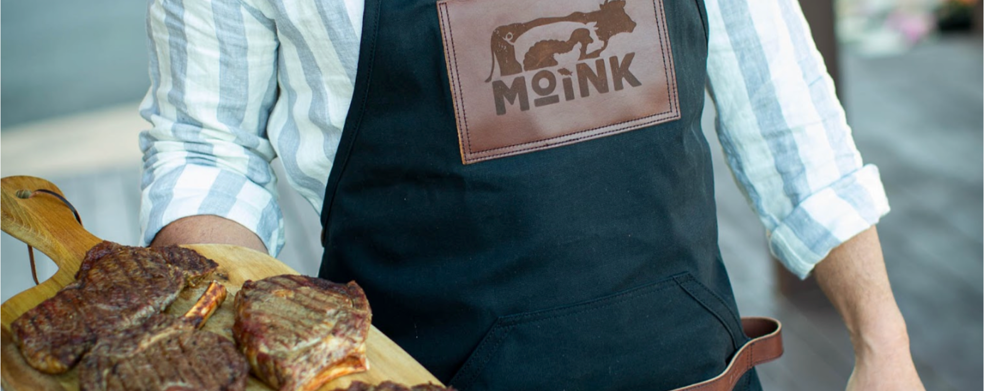 Person wearing a black apron with a 'Moink' logo, holding a wooden board with steaks.