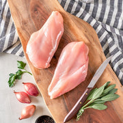 Two boneless skinless chicken breasts on a wooden cutting board with a knife, garlic cloves, and parsley leaves on the side.
