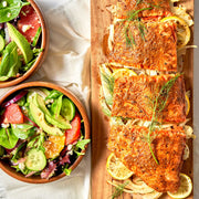 Salmon fillets with herbs and lemons on a wooden board, next to a salad in a bowl.