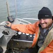 Fisherman smiling while operating a boat, wearing an orange jacket and black beanie.