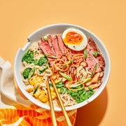 Bowl of ramen with beef, noodles, and vegetables on an orange background.