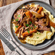 Braised meat with mushrooms and carrots over mashed potatoes on a wooden table.
