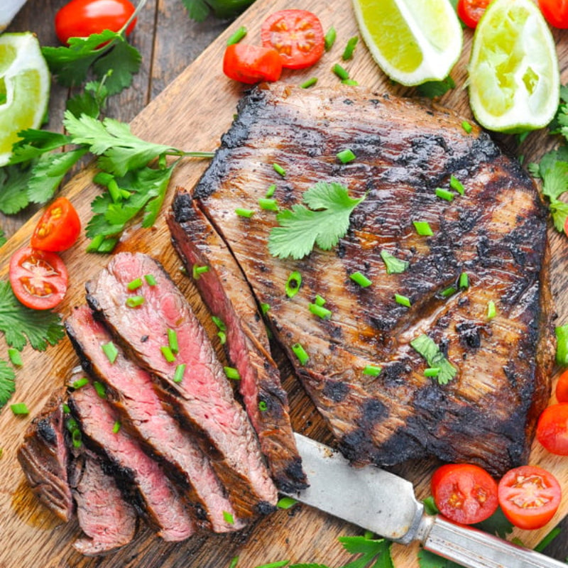 Grilled steak with slices cut, surrounded by tomatoes and limes on a wooden board.