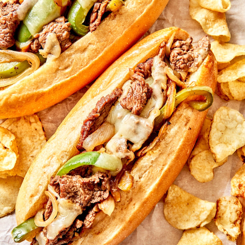 Two philly cheese steaks with chips on a wooden board.