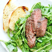 Grilled steak on arugula with toasted bread on a white plate.