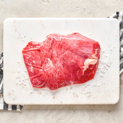 A raw flank steak on a white cutting board with a striped kitchen towel in the background.