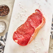A raw NY Strip steak on a white cutting board, surrounded by salt, pepper, and a striped napkin.
