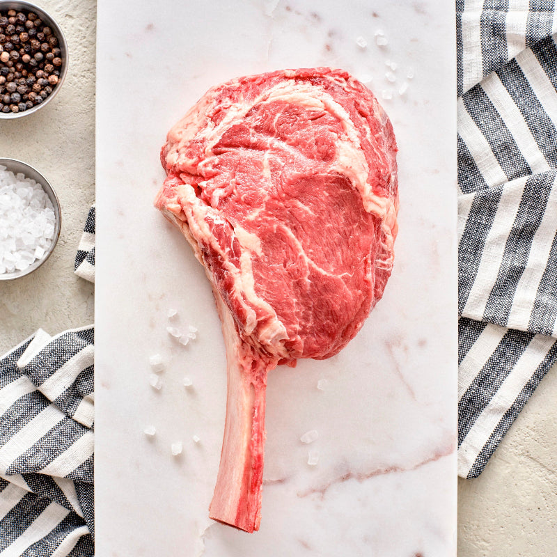 A bone-in rib-eye steak on a white marble surface with a striped napkin, salt, and pepper.
