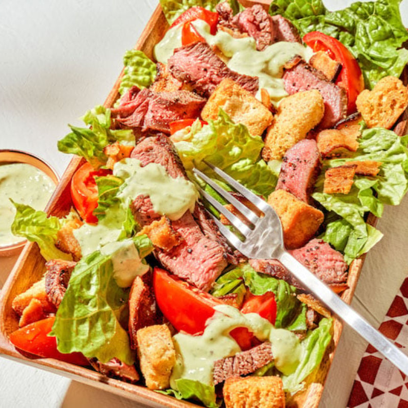 Salad with meat, vegetables, and croutons in a tray with a fork and dressing.