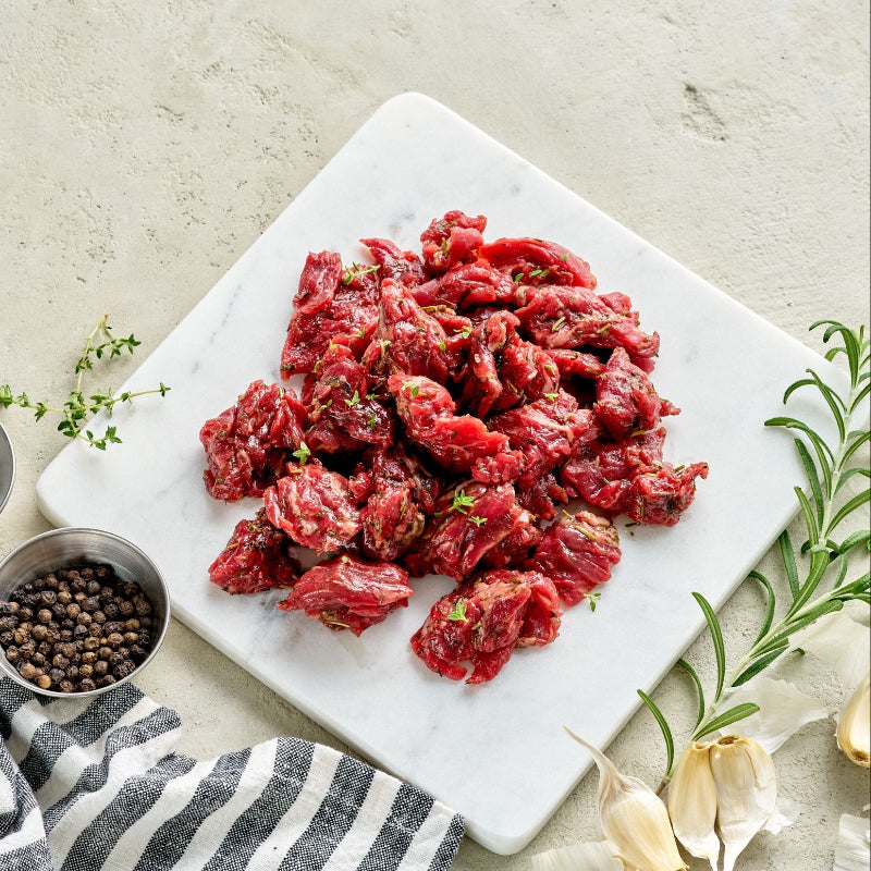 Raw marinated beef pieces on a marble cutting board with herbs and spices.