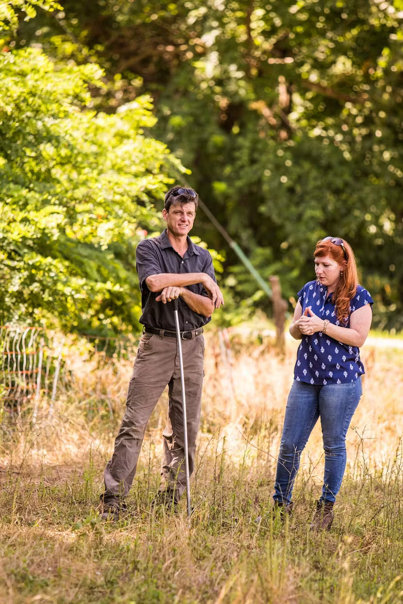 Lucinda and a farmer standing in a grassy field with trees in the background