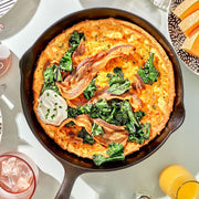 Skillet with frittata topped with bacon and spinach on a table with drinks and plates.