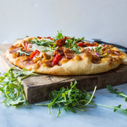 Bacon pizza with arugula on a wooden cutting board.