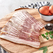 Sliced bacon on a wooden cutting board with a small bowl of eggs and some herbs in the background.