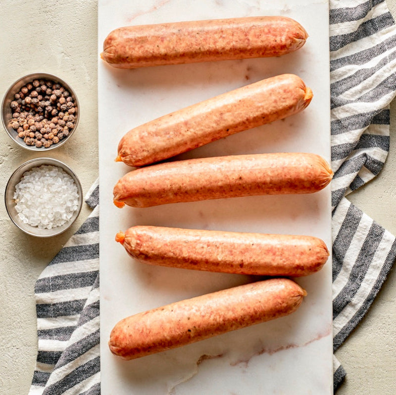 Raw pork brats on a marble surface with a side of spices and salt, striped napkin to the side.