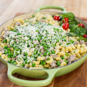 Green skillet with pasta, pork, vegetables, and cheese on a wooden surface