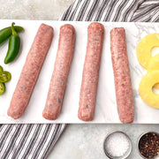 Raw pineapple jalapeno brats laid out on a white surface, surrounded by ingredients such as jalapeno peppers, pineapple rings, and seasonings.
