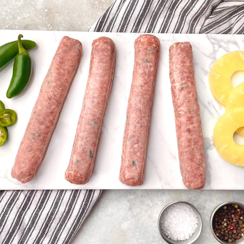 Raw pineapple jalapeno brats laid out on a white surface, surrounded by ingredients such as jalapeno peppers, pineapple rings, and seasonings.