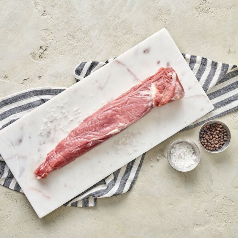 A raw pork tenderloin on a white marble cutting board, flanked by a small bowl of salt and a dish of ground pepper, with a striped napkin in the background.