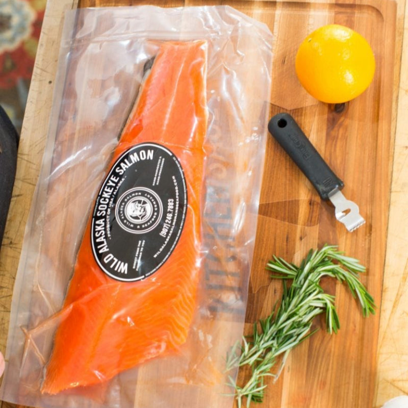A large Sockeye salmon filet is packaged and displayed on a wooden cutting board, accompanied by a lemon and some fresh herbs.