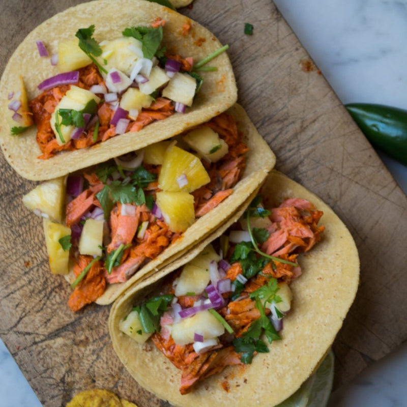 Three salmon tacos with pineapple and onions on a wooden board.