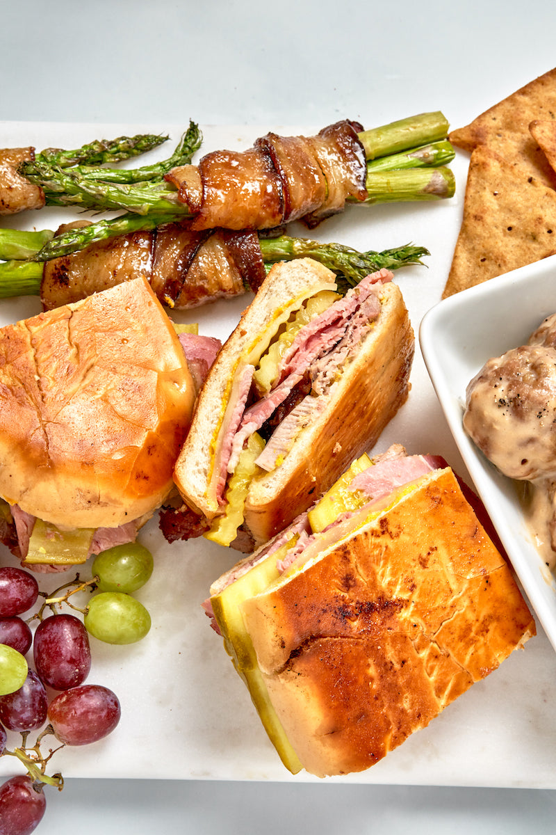 Assorted food items including sandwiches, asparagus, grapes, and bread on a white plate.
